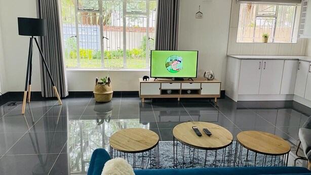 A modern living room with a TV, two round coffee tables, a blue sofa, and a kitchen area with cabinets. - Bulawayo Bradfield Retreat - 3 Bed House (Bulawayo)