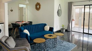 A modern living room with a blue sofa, a grey armchair, and two small round tables. A large mirror hangs on the wall, and there is a potted plant on the floor. - Bulawayo Bradfield Retreat - 3 Bed House (Bulawayo)