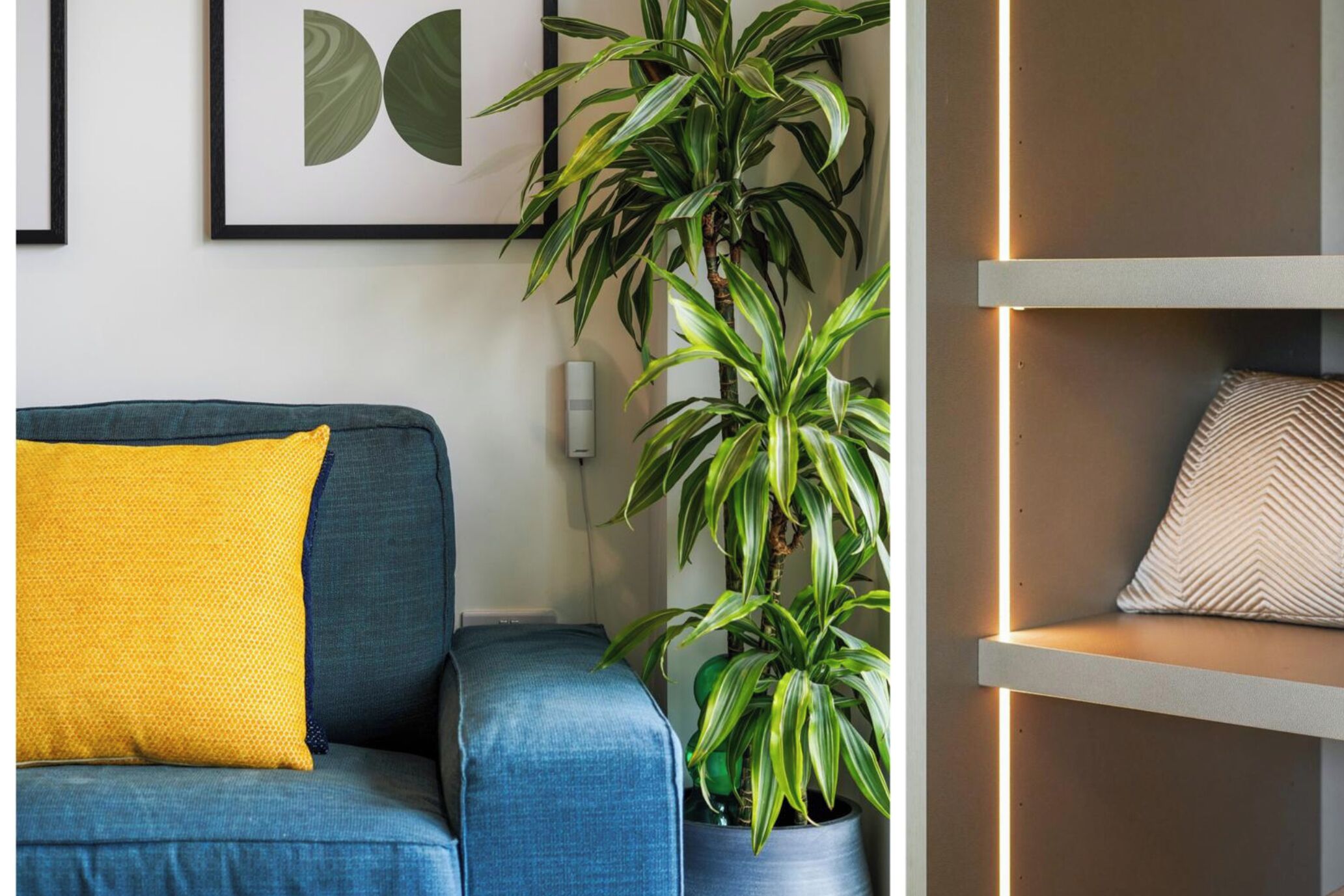 En stue med en blå sofa, en gul pude, en potteplante og en vægmonteret hylde med en pudepude.