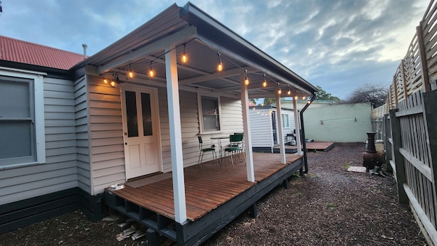 Une maison dotée d’une terrasse en bois, d’un porche illuminé de guirlandes lumineuses et d’un petit espace extérieur aménagé pour s’asseoir.