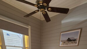 Une chambre avec un plancher en bois, un lit recouvert d’une couvre-lit beige et blanche, un ventilateur de plafond, une fenêtre avec des stores et un tableau encadré accroché au mur.