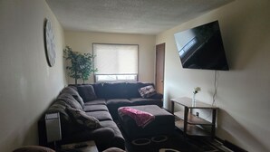 Een woonkamer met een hoekbank, een flatscreen televisie aan de muur, een potplant en een raam met jaloezieën.