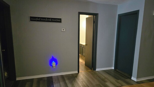 Een hal met een blauw verlicht wandlamp, een deur die naar een andere kamer leidt, en een houten tafel op de voorgrond.