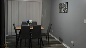 Een eethoek met een tafel, stoelen, een raam met zonwering en een aan de muur hangend schilderij.