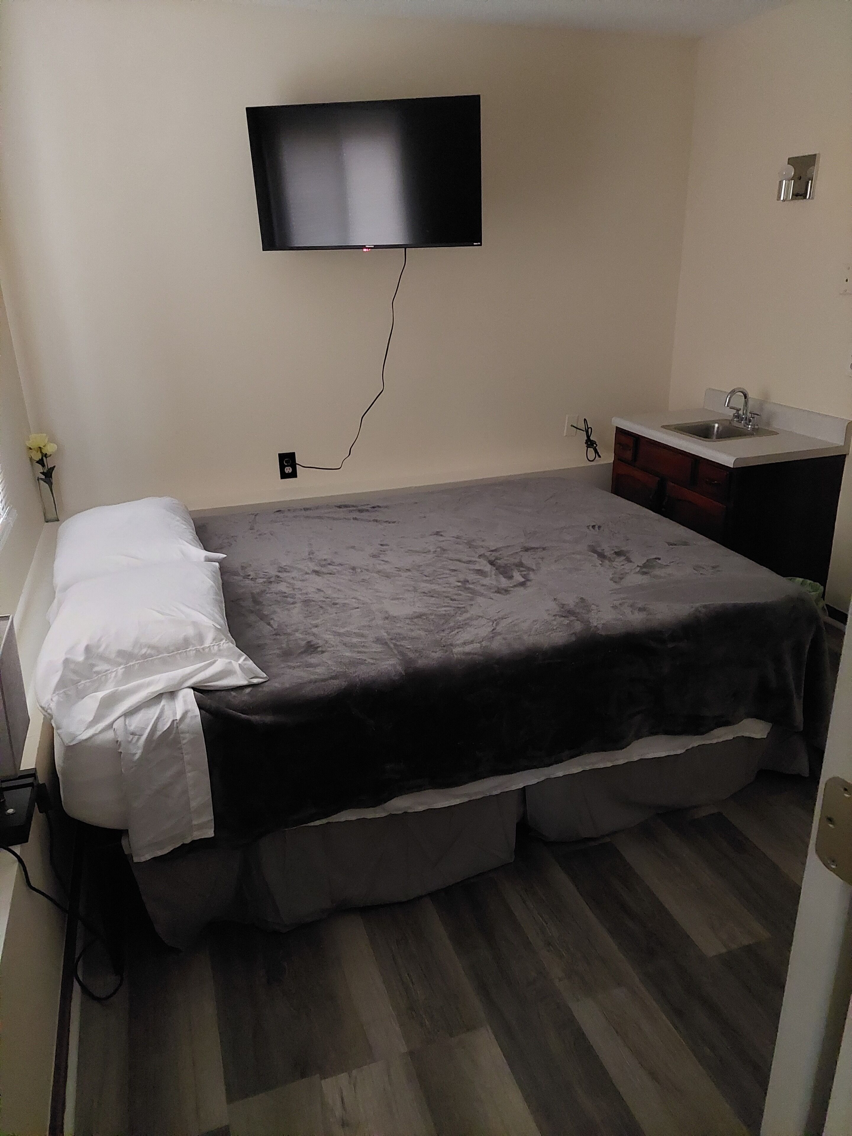 A bedroom with a bed, a flat-screen TV mounted on the wall, a small sink, and a wooden floor.