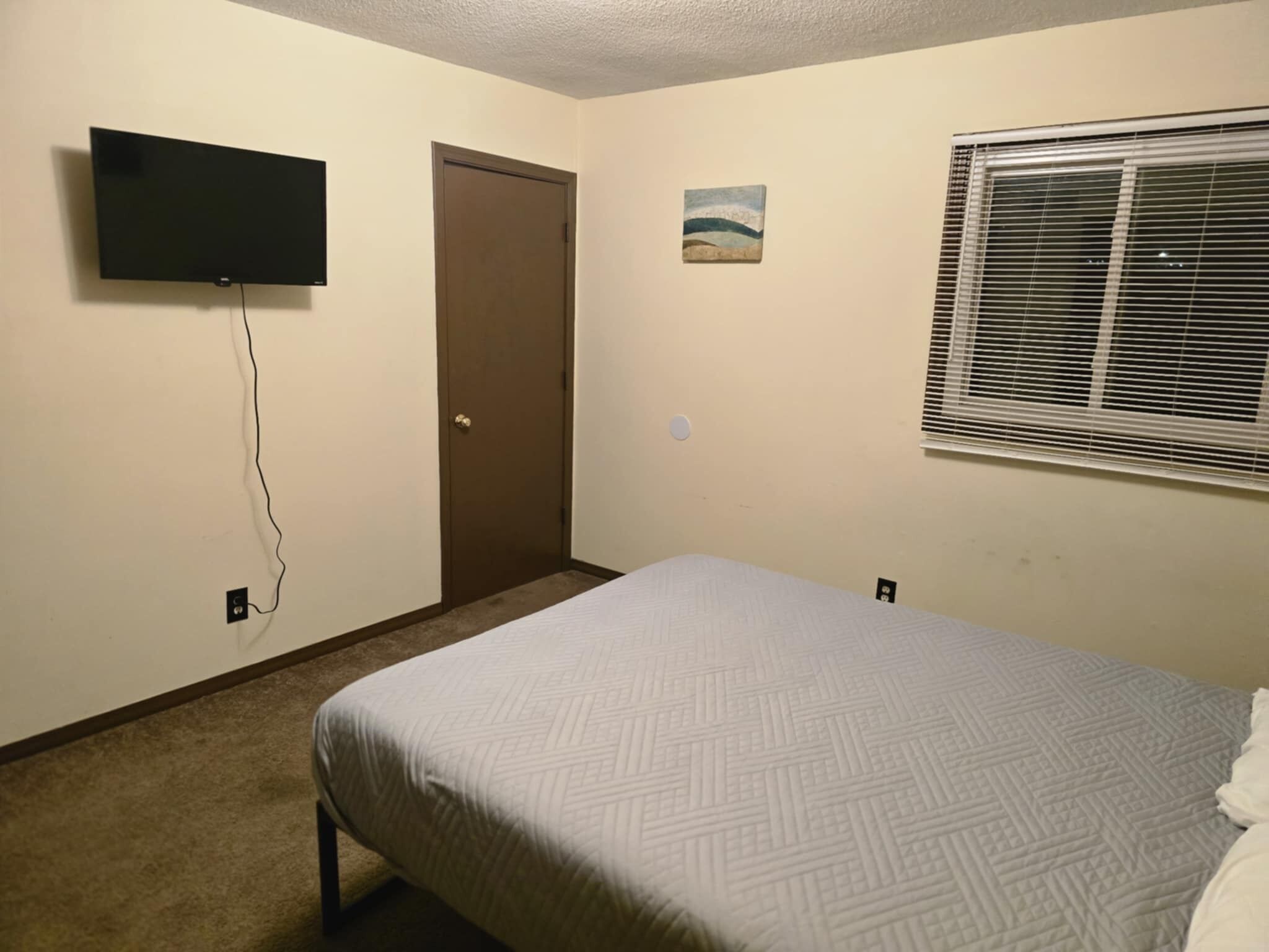 A bedroom with a bed, a television mounted on the wall, a door, a small painting, and a window with blinds.