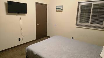 A bedroom with a bed, a television mounted on the wall, a door, a small painting, and a window with blinds.