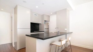 A modern kitchen with white cabinetry, a black countertop, and stainless steel appliances. There are three bar stools and a sink. - 304 Arena Newcastle Beach (Newcastle, New South Wales, Australia)