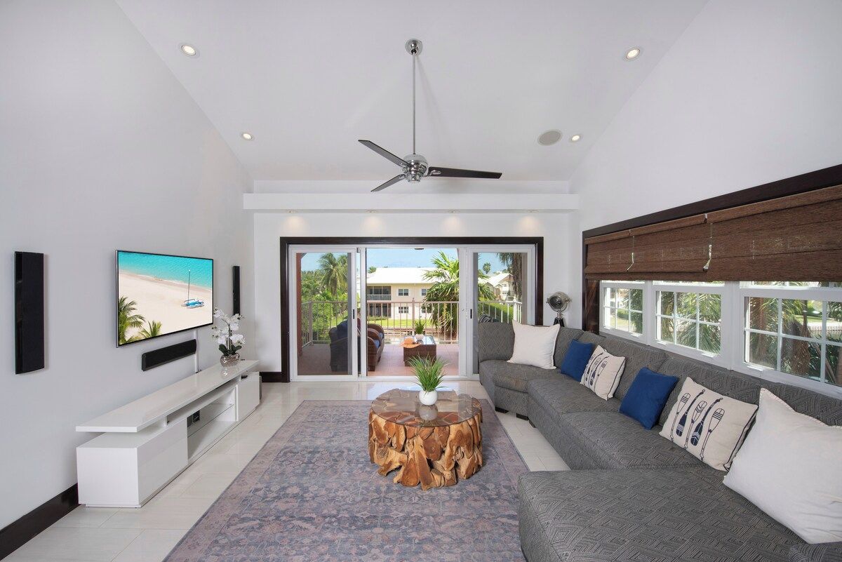 Ein modernes Wohnzimmer mit einem Flachbildfernseher, einem Deckenventilator, einer Couch mit gemusterten Kissen und einem großen Fenster mit Blick ins Freie.