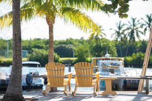 A wooden deck with two Adirondack chairs and a table, overlooking a marina with boats and palm trees.