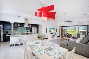 A modern kitchen with a central island, a dining area with a patterned tablecloth, and a living area with a sofa and a large window. - Canal Front 3BR Condo on 7 Mile Beach - Britannia 634 (Brittania)