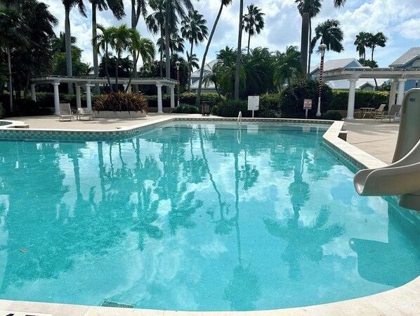 A clear swimming pool with a slide, surrounded by palm trees and a covered seating area.
