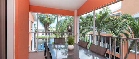 Een balkon met een glazen tafel en stoelen, een bloempot en uitzicht op palmbomen en een zwembad.