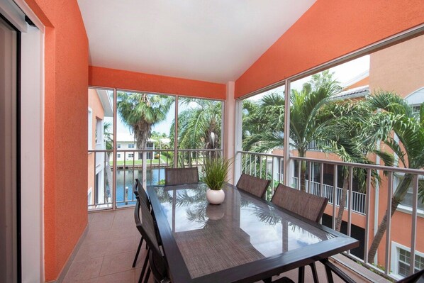A balcony with a glass table and chairs, a potted plant, and a view of palm trees and a pool.