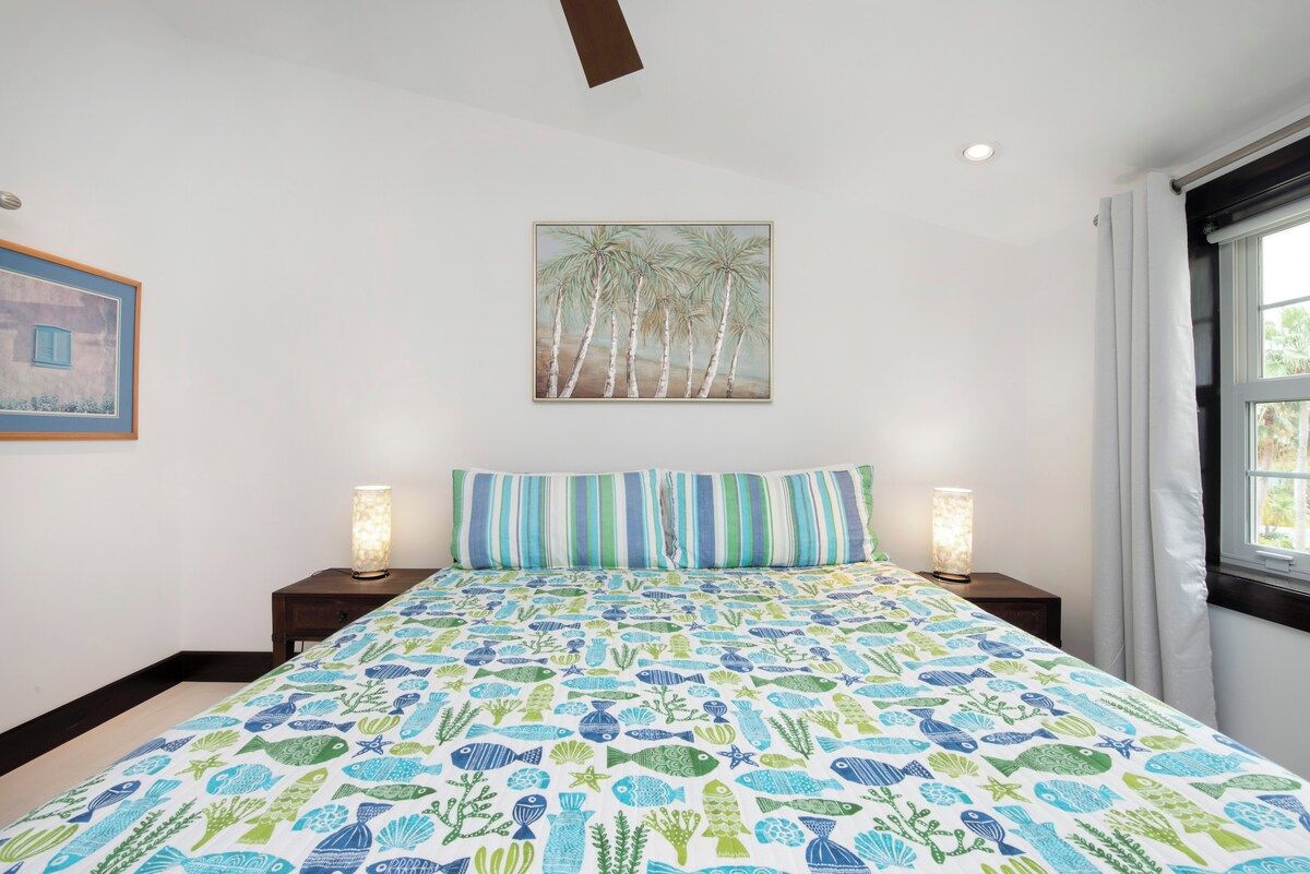 A bedroom with a bed, striped pillows, a wall painting, two bedside lamps, and a window with curtains.