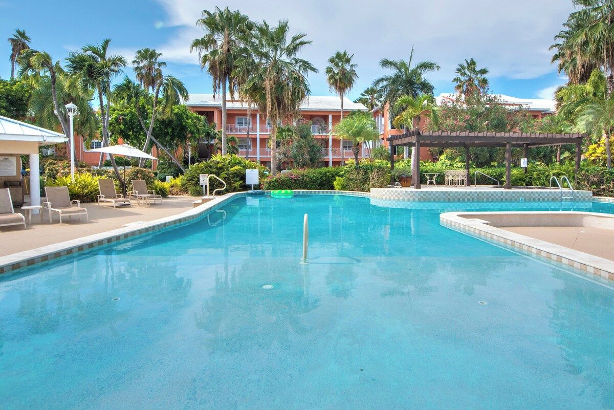 A swimming pool with lounge chairs and umbrellas, surrounded by palm trees and a building with balconies.