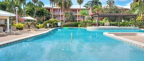 A swimming pool with lounge chairs and umbrellas, surrounded by palm trees and a building with balconies.