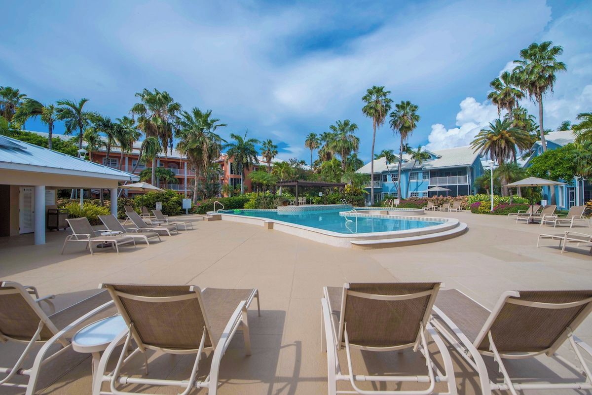 A resort pool area with lounge chairs, palm trees, and a clear sky.
