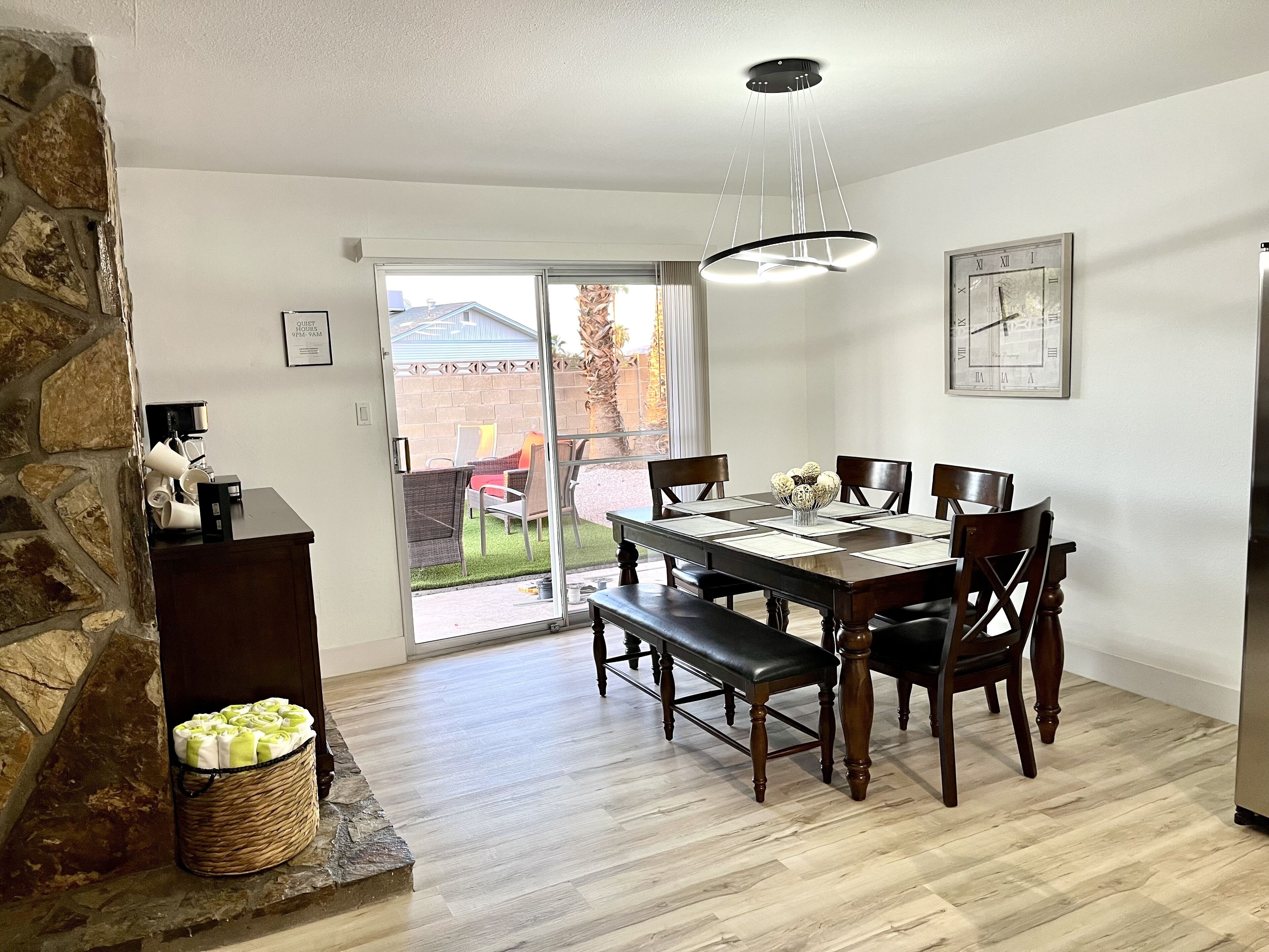 A dining area with a wooden table, chairs, a bench, a stone wall, a basket of towels, and a clock on the wall.