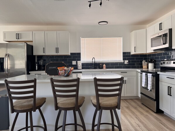 A modern kitchen with white cabinets, a black backsplash, and stainless steel appliances. There are three bar stools and a welcome sign on the counter. - HEATED Pool, Spacious home 7 MIN to airport* (Las Vegas)