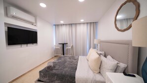 Een moderne slaapkamer met een groot bed, een flatscreen-televisie aan de muur, een klein tafeltje met twee stoelen en een luchtverfrisser aan het plafond.