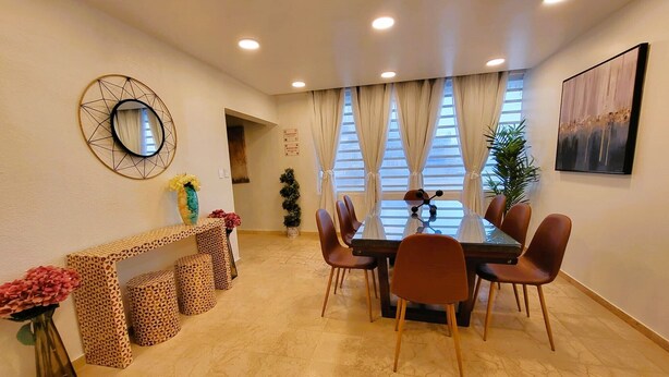 Een eetgedeelte met een ronde tafel, zes stoelen, een decoratieve spiegel, een vaas met bloemen en een console tafel met een patroon.