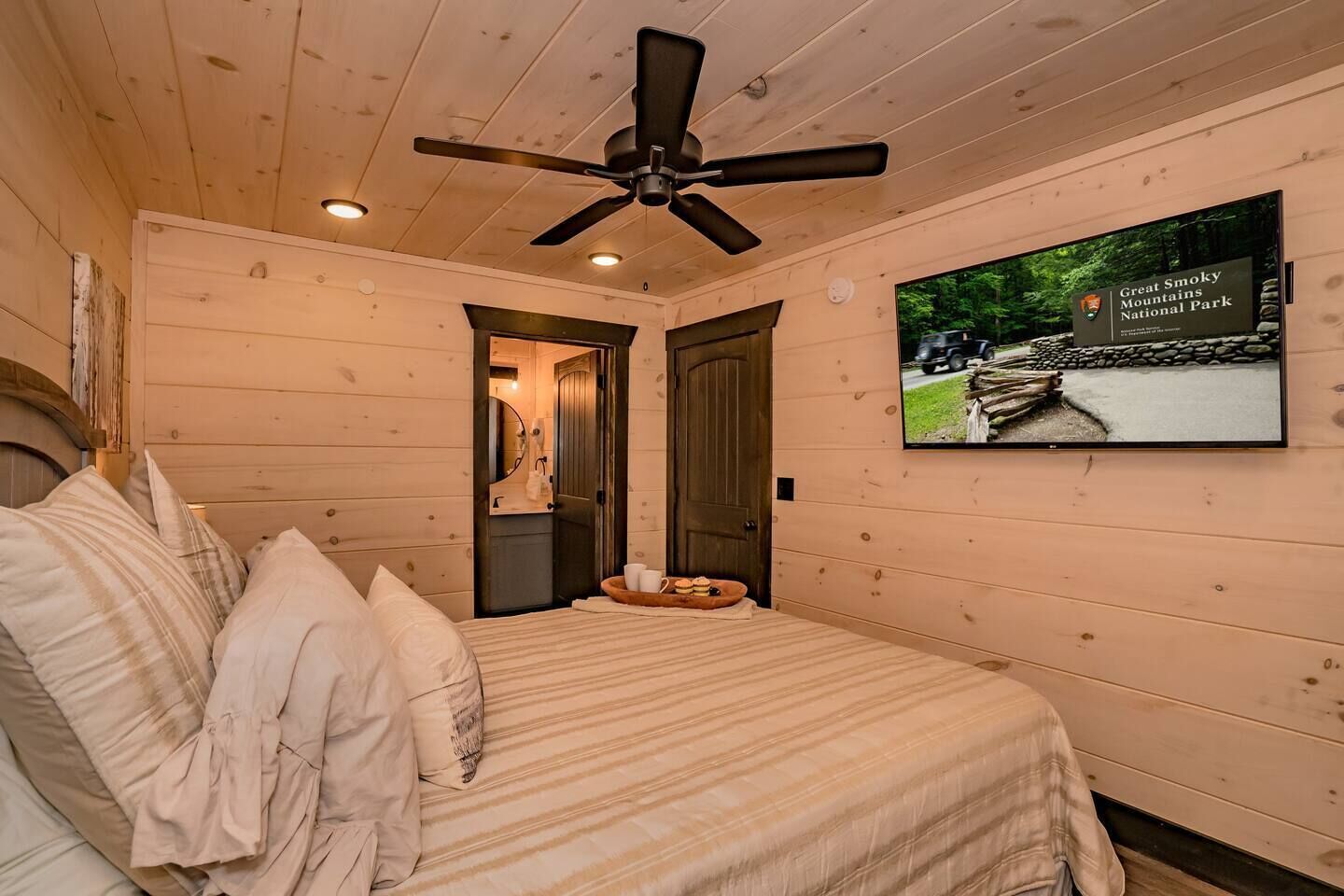 A bedroom with a bed, a ceiling fan, a television displaying Great Smoky Mountains National Park, and a wooden door leading to another room.