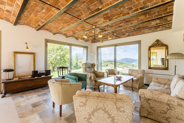 A spacious living room with a high ceiling, large windows offering a view of the countryside, and a mix of upholstered furniture.