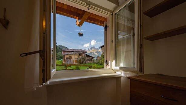 Ein Zimmer mit Fenster und Blick auf eine Berglandschaft, Holzbalkendecke und ein Holzkasten.