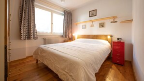 Ein Schlafzimmer mit einem hölzernen Bett, einer roten Kommode, einem Fenster mit gemusterten Vorhängen und einem an der Wand befestigten Regal mit Bildern.