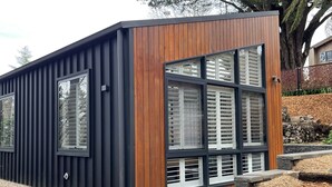 A modern building with a combination of dark siding and wooden paneling, large windows with white shutters, and a landscaped garden area with mulch and gravel. - Contemporary, Stylish One Bedroom Studio. (Katoomba)