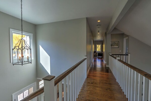 A long, narrow hallway with wooden floors and white railings, leading to a room with a window and a chandelier.