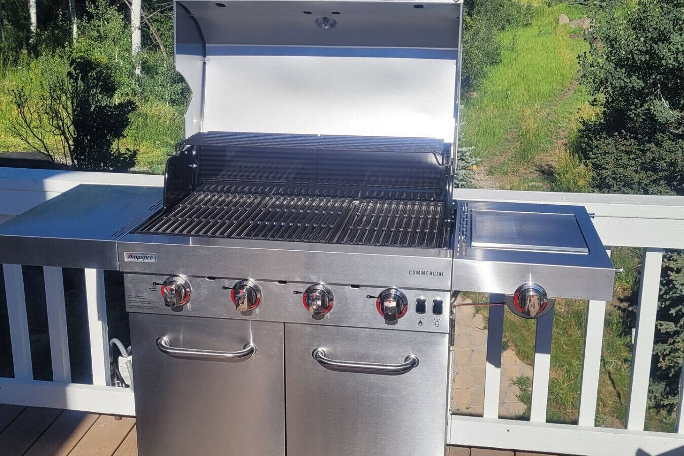 En professionel grill med fire brændere og en ekstra sidebrænder, placeret på en terrasse med gelænder i hvid.