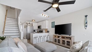A modern living room with a sofa, ottoman, and a TV on a wooden cabinet. A kitchen with white cabinets and a bar area is visible in the background.