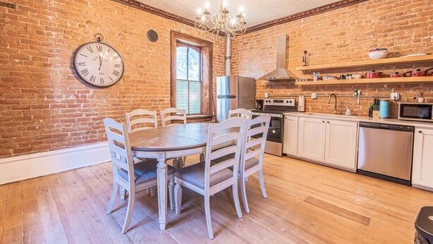 A kitchen with a brick wall, wooden dining table, white cabinets, stainless steel appliances, and a clock on the wall.