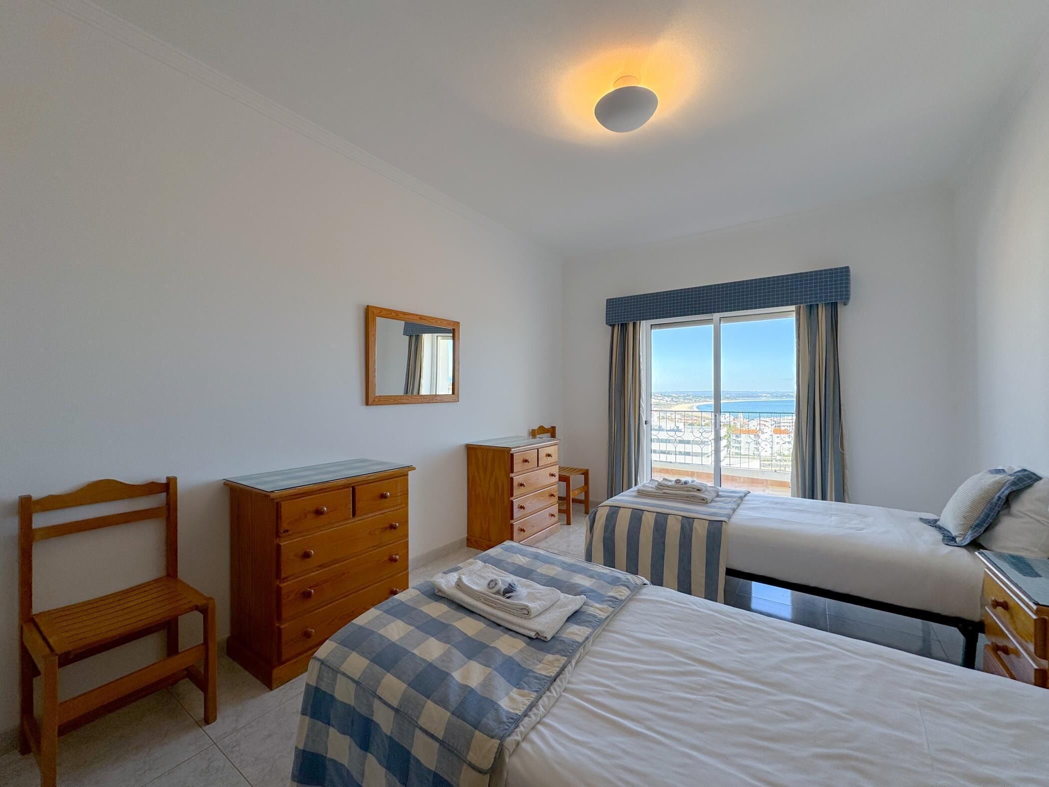 A bedroom with two beds, a wooden chair, a dresser, and a window with a view of the sea.