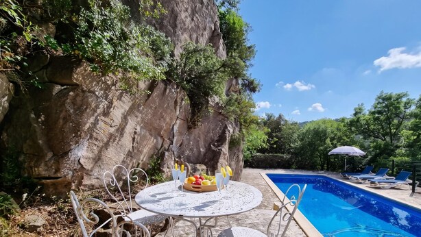 An outdoor pool area with a table and chairs, surrounded by greenery and a rocky cliff.