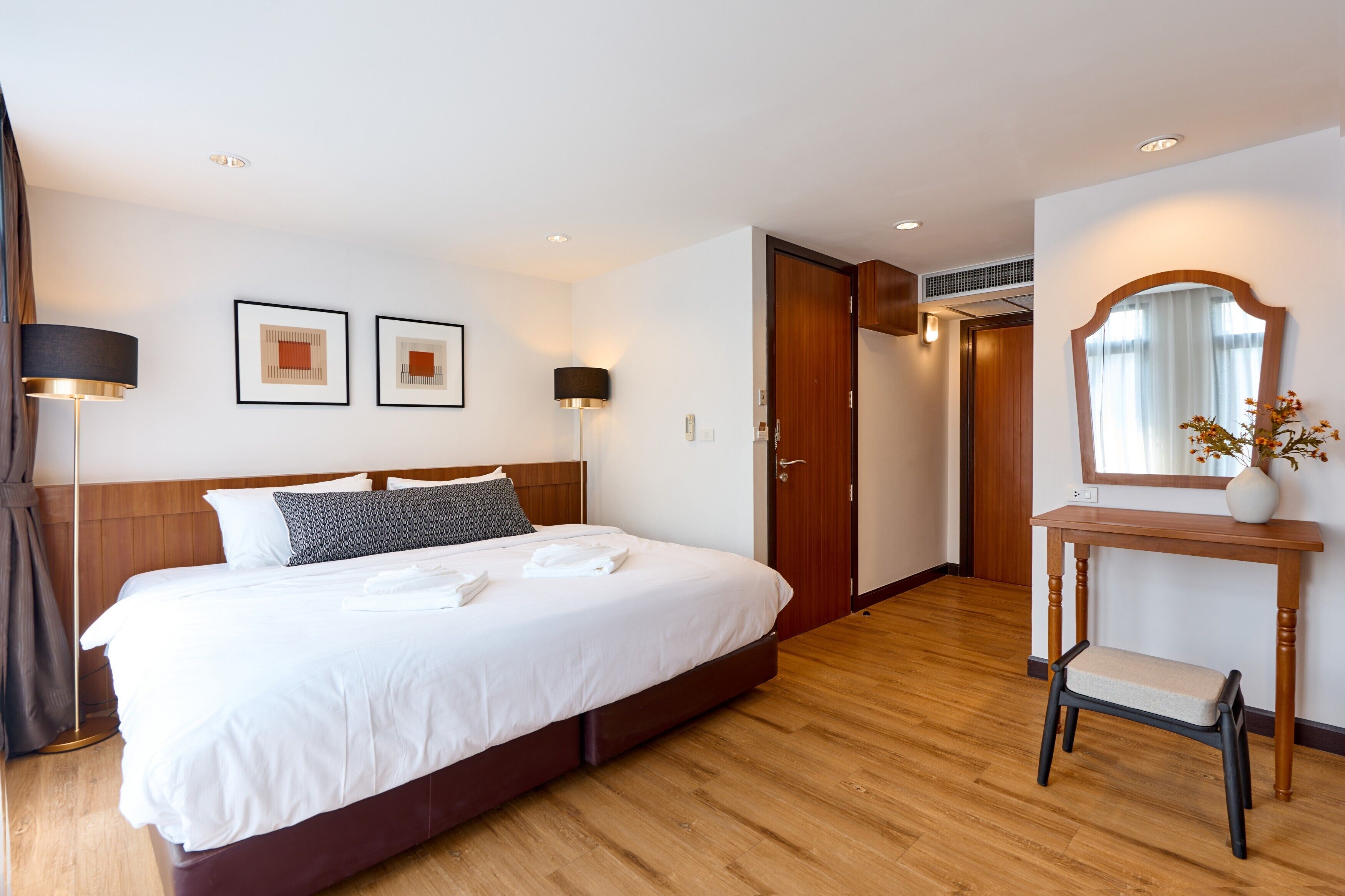 Une chambre d’hôtel avec un grand lit, des lampes de chevet, une tête de lit en bois, un miroir et une petite table sur laquelle est posé un vase.