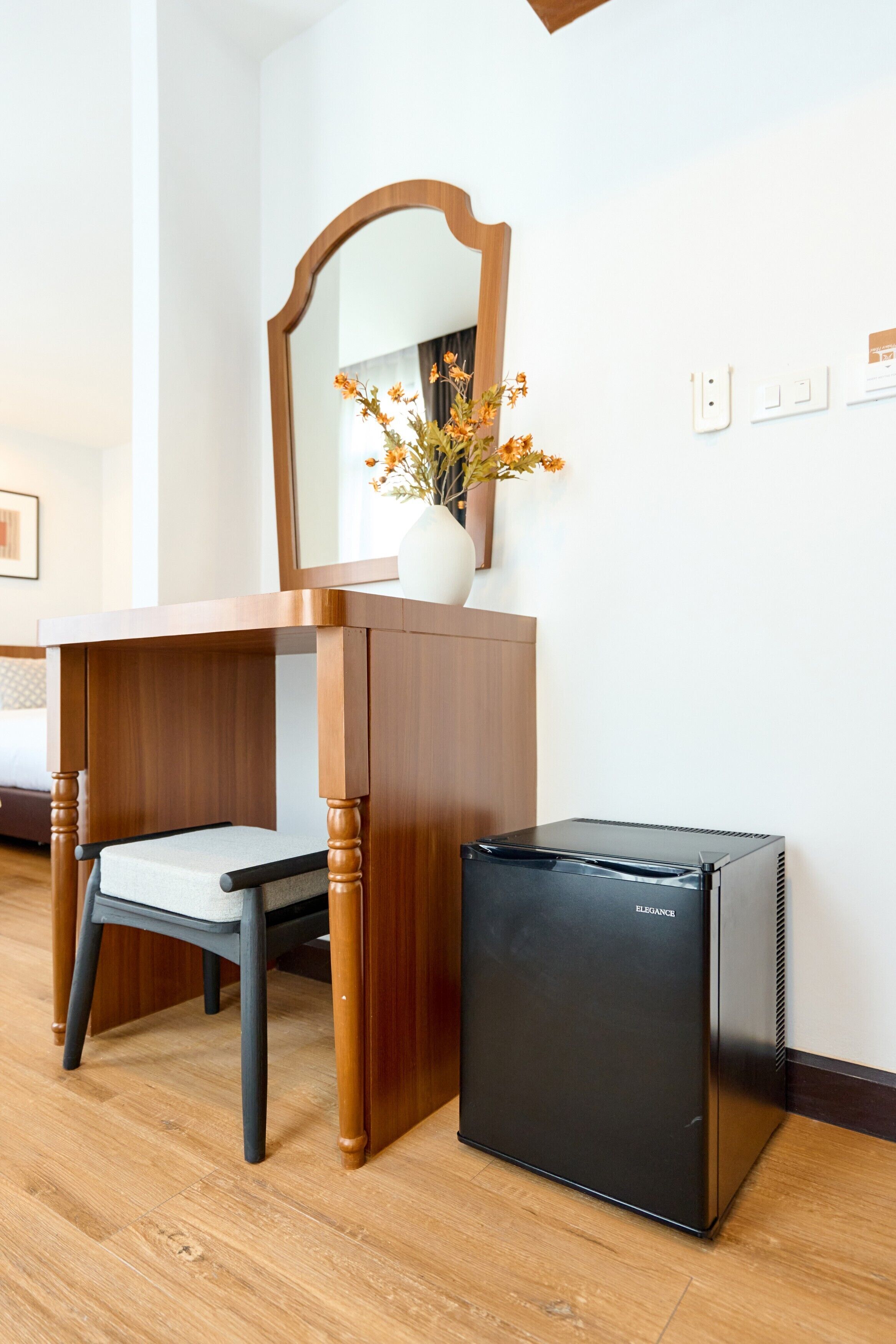 A compact room with a wooden desk, a chair, a small black refrigerator, a mirror, and a vase with flowers on the wall.