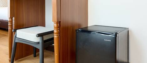 Une pièce de taille modeste, avec un bureau en bois, une chaise, un petit réfrigérateur noir, un miroir et un vase avec des fleurs accroché au mur.