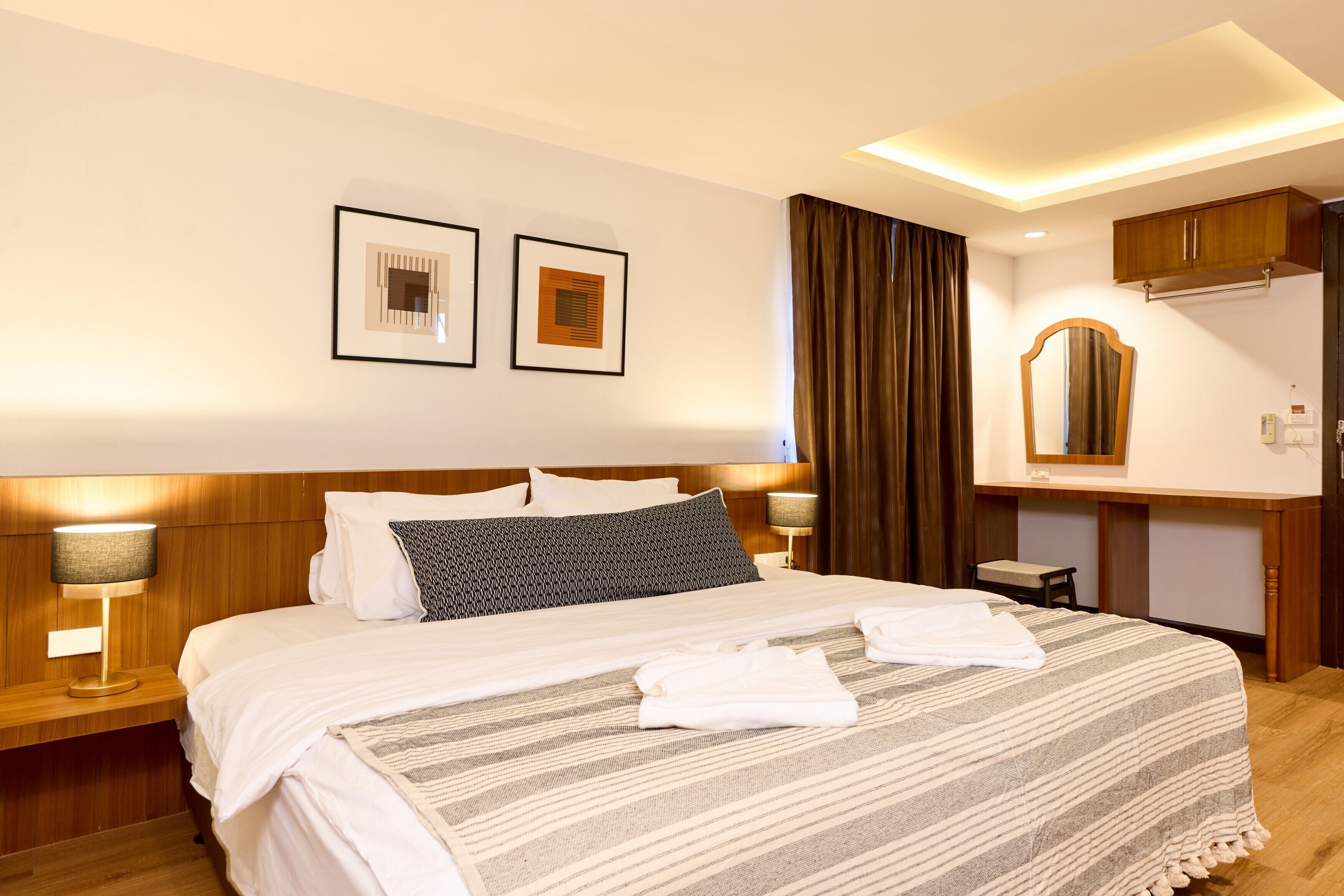 A hotel room with a large bed, two framed pictures on the wall, a wooden headboard, a bedside table with a lamp, a mirror, and a wooden cabinet.