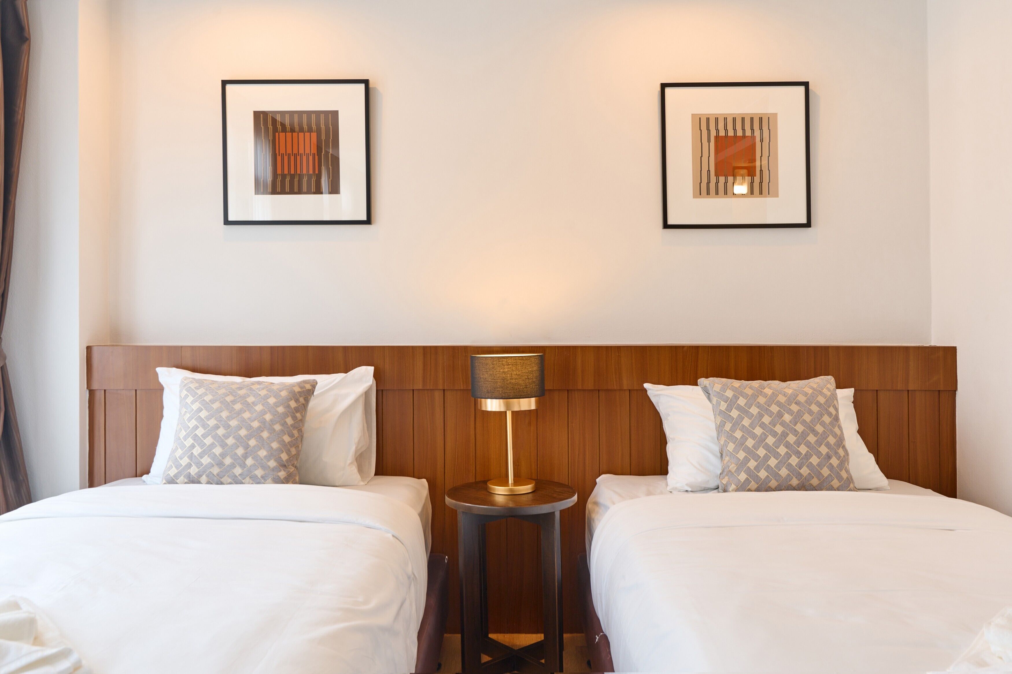 A hotel room with two beds, a wooden headboard, a round table with a lamp, and two framed artworks on the wall.