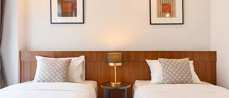A hotel room with two beds, a wooden headboard, a round table with a lamp, and two framed artworks on the wall.