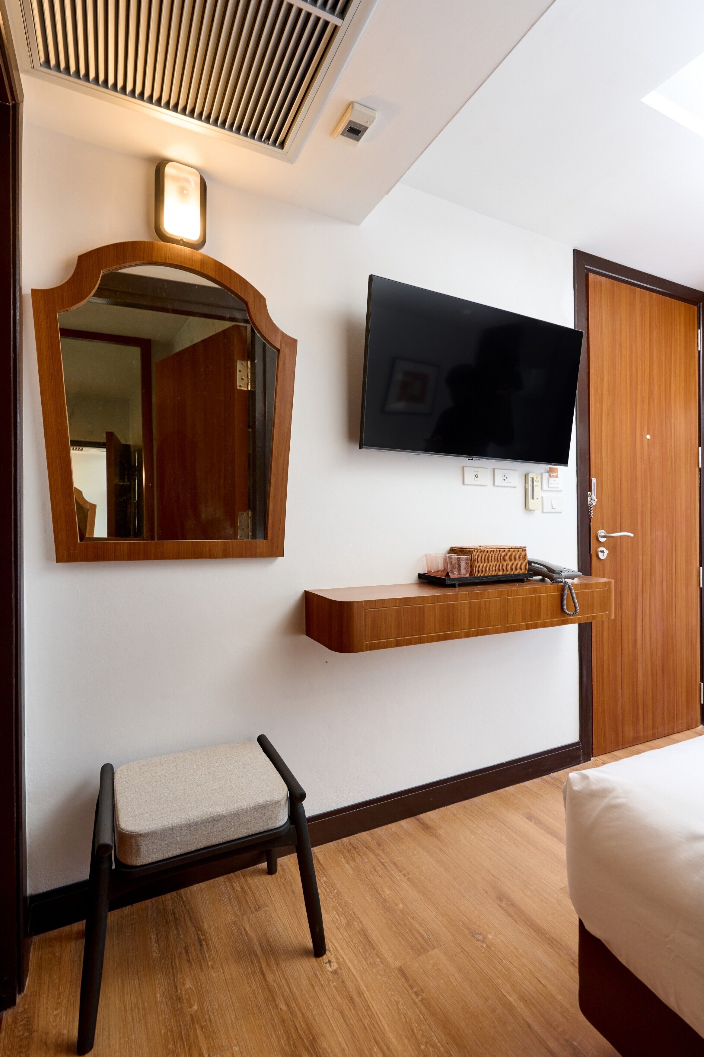 A hotel room with a wooden floor, a bed, a television mounted on the wall, a wooden bench, a mirror, and a shelf with a lamp.