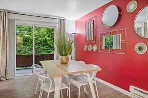 A room with a red wall, a wooden table, white chairs, and a large window with a view of greenery.