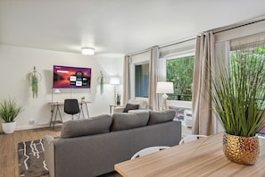 Een moderne woonkamer met een grijze bank, een houten eettafel en een televisie aan de muur waarop diverse streamingdiensten worden weergegeven.