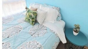 A bedroom with a blue wall, a bed with patterned bedding, two wall art pieces, a window with sheer curtains, and a small potted plant on a round table beside the bed. - Beautiful apartment steps from the subway (Santiago)
