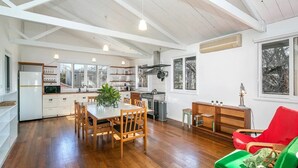 A spacious kitchen with wooden flooring, a dining area with a table and chairs, and a bright, airy living room with a red chair and a green ottoman. - Marguerite House by Free Trade Co (Braidwood)