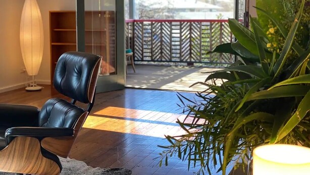 A modern living room with a leather chair, a floor lamp, and a large plant. A glass door leads to an outdoor area with a view of trees and a fence. - Marguerite House by Free Trade Co (Braidwood)