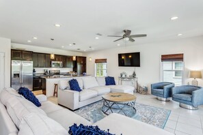 A spacious living area with a white sofa, blue throw pillows, a coffee table, and two blue armchairs. A kitchen with dark cabinets and stainless steel appliances is visible in the background.